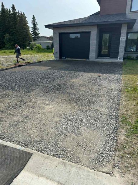 A man walks along a gravel driveway, surrounded by greenery and a clear blue sky.